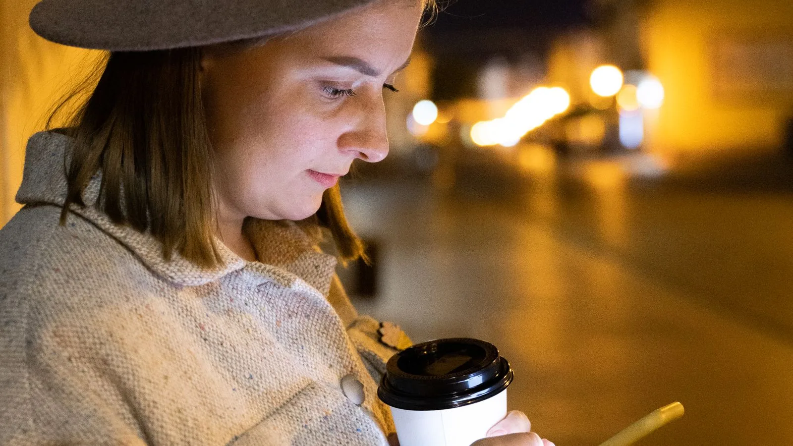A traveller checking her phone over a coffee, warm city lights blurred behind her